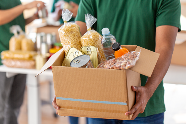 Volunteer in green shirts holding a cardboard box of non-perishable food supplies with volunteers packing food in background.