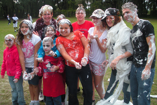 Group of smiling teens and and adults with shaving cream and toilet paper all over them outside.