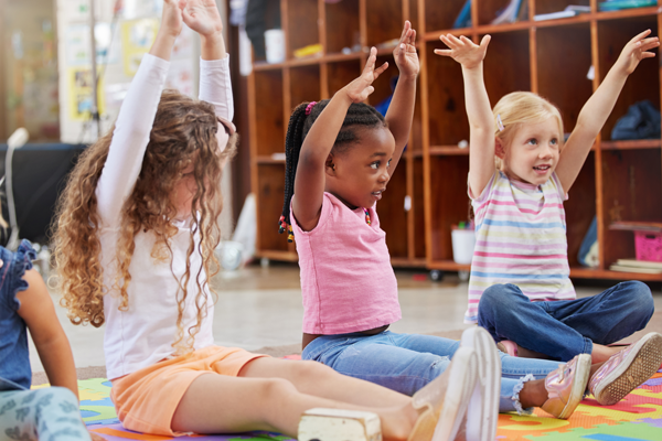 Three diverse little girls, happy and stretching with arms up, sitting on floor in colorful daycare center.