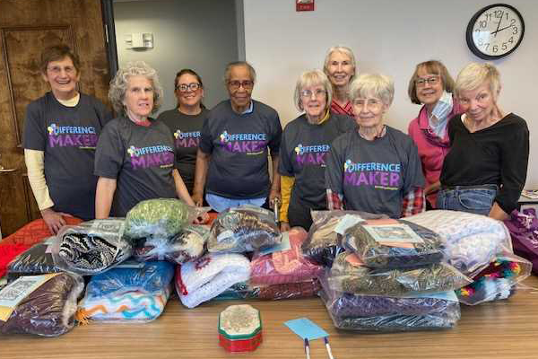 photo of Large group of happy women with Difference Maker t-shirts standing behind a table filled with blankets.